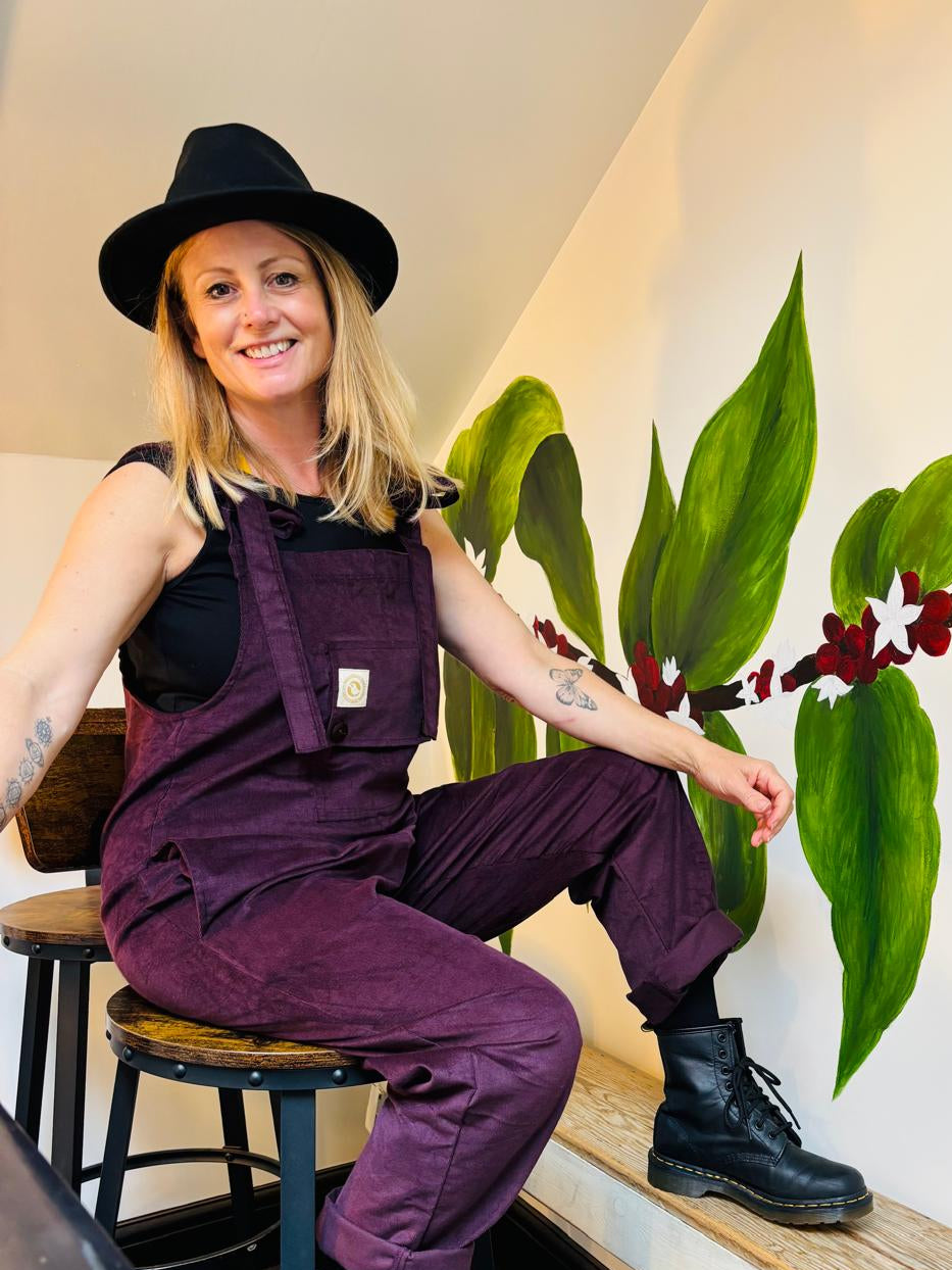 Person wearing purple overalls sitting on a stool next to a wall with green plant art.