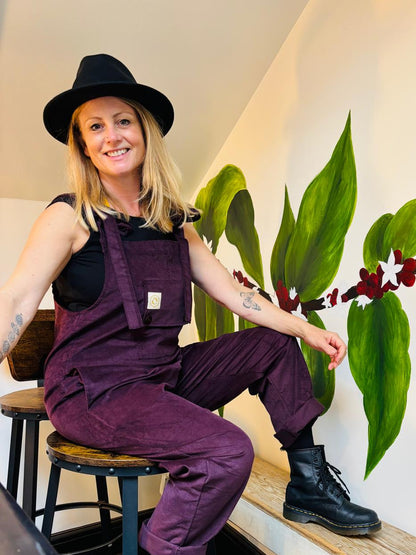 Person wearing purple overalls sitting on a stool next to a wall with green plant art.
